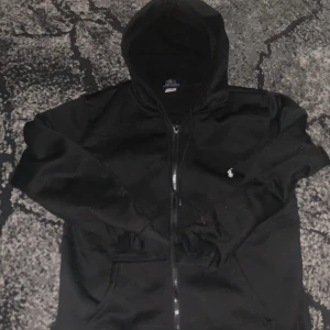 Svart hoodie från Polo Ralph Lauren - Svart hoodie från Polo Ralph Lauren med klassisk vit logga broderad på bröstet. Tröjan har huva med snörning, dragkedja framtill och långa ärmar. Materialet är mjuk bomull och passformen är normal. Perfekt för chill dagar och streetwear.