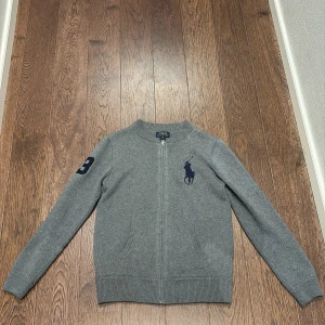 Grå stickad kofta från Polo Ralph Lauren - Snygg grå stickad kofta från Polo Ralph Lauren med dragkedja framtill, ribbade muddar och två fickor. Mörkblå Polo-logga på bröstet och en stor siffra 3 på ärmen ger en sportig vibe. Perfekt för dig som gillar klassisk stil med en twist.