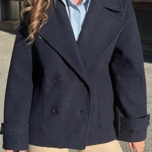 Mörkblå skepparkavaj med bred krage - Säljer en stilren mörkblå skepparkavaj med bred krage och dubbelknäppning framtill. Jackan har långa ärmar med dekorativa knappar vid mudden och en klassisk, rak passform. Perfekt för dig som gillar tidlös och snygg stil.