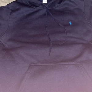 Mörkblå hoodie från Polo Ralph Lauren - Snygg mörkblå hoodie från Polo Ralph Lauren med klassisk broderad logga i blått på bröstet. Tröjan har huva med snörning och en stor magficka framtill. Perfekt för dig som gillar stilren streetwear och vill ha något bekvämt och coolt.