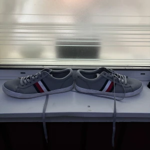 Grå sneakers Tommy Hilfiger - Snygga grå sneakers från Tommy Hilfiger med klassiska ränder i rött, vitt och marinblått på sidan. Skorna har rund tå, platt vit sula och snörning. Perfekta för dig som gillar stilrena och sportiga skor med en touch av färg.