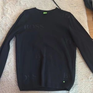 Svart stickad tröja från Hugo Boss - Svart stickad tröja från Hugo Boss med diskret BOSS-logga ton-i-ton på framsidan och en grön etikett vid nederkanten. Tröjan har rund hals, ribbade muddar och långärmad passform. Perfekt för dig som gillar stilrena och klassiska plagg med en modern touch.