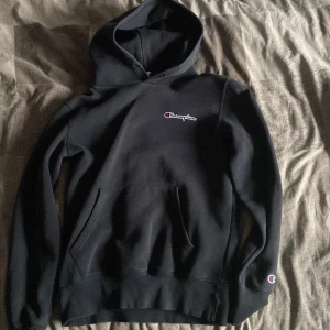 Svart Champion hoodie med huva - Svart hoodie från Champion i storlek small. Klassisk design med broderad Champion-logga på bröstet och ärmen, stor huva och magficka. Mjuk och skön bomullsmix, perfekt för chill eller streetwear. Snygg passform och långärmad modell.