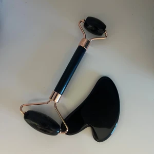 Svart ansiktsroller och gua sha-set - Snyggt set med svart ansiktsroller och matchande gua sha-sten i polerad sten. Rollern har detaljer i roséguld och två rullar för ansiktsmassage. Perfekt för att boosta hudvårdsrutinen och ge ansiktet extra glow.