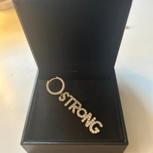 Glittrig örhänge med texten STRONG - Snyggt örhänge i silverfärg med hängande bokstäver som bildar ordet STRONG, täckta av glittrande stenar. Perfekt statement piece för dig som vill sticka ut och visa elegans 