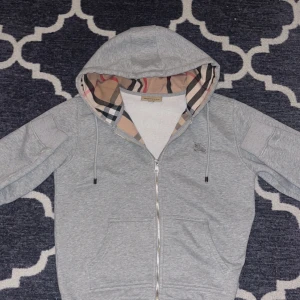 Grå Burberry hoodie med dragkedja - Snygg grå hoodie från Burberry med klassiskt rutigt foder i huvan och insidan. Tröjan har dragkedja framtill, snörning vid huvan och två fickor. Materialet är mjuk bomull med lite elastan för extra komfort. Perfekt för dig som gillar stilrena och lyxiga detaljer.