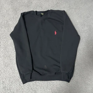 Svart sweatshirt från Polo Ralph Lauren - Svart sweatshirt från Polo Ralph Lauren med klassisk röd broderad logga på bröstet. Tröjan har rund hals, ribbade muddar och är tillverkad i mjuk bomull. Perfekt för dig som gillar stilrena och enkla plagg med en touch av lyx.