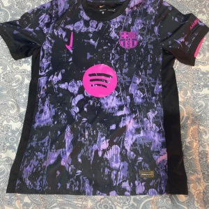 Barcelona Lamine Yamal Nike fotbollströja M - Säljer en Barcelona fotbollströja med Lamine Yamal och nummer 19 på ryggen. Tröjan har coolt svart och lila mönster med rosa detaljer, Nike-logga och Spotify sponsor. Tillverkad i lätt Dri-Fit material som andas, perfekt för match eller träning.