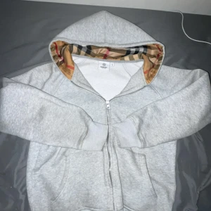 Grå Burberry hoodie med rutig huva - Säljer en grå hoodie från Burberry i storlek xxs-xsTröjan har dragkedja framtill, klassisk huva med ikoniskt Burberry-rutigt foder i beige, svart, vit och röd. Tillverkad i mjuk bomull, perfekt för chill dagar. Stora fickor framtill och ribbade muddar vid ärmslut och nederkant.