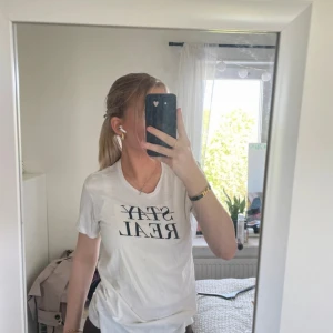 Vit t-shirt med tryck från FSBN - Vit t-shirt från FSBN med svart texttryck 'STAY REAL' på bröstet. Klassisk rund halsringning och korta ärmar. Skön och lätt i bomull, perfekt för en chill och enkel stil.