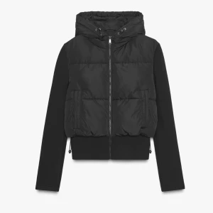 Svart pufferjacka med huva - Svart pufferjacka med huva och snörning, dragkedja framtill och långa ärmar. Jackan har en quiltad look på kroppen och släta ärmar, perfekt för kyliga dagar. Snygg och enkel design som funkar till många olika stilar.