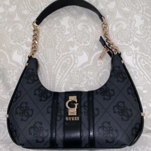 Svart handväska från Guess med kedja -  ALDRIG ANVÄND.Snygg svart handväska från Guess med guldiga kedjedetaljer och logotyp framtill. Väskan har ett mönstrat tyg med Guess-emblem och svarta skinnpaneler. Dragkedja upptill och axelrem med både kedja och skinn. Perfekt storlek för det viktigaste. Dust bag följer med.