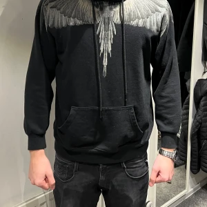 Marcelo burlon hoodie - Svart hoodie från Marcelo Burlon County of Milan i storlek M. Tröjan har ett grått vingmönster över axlar och bröst, huva med snörning och stor magficka. Tillverkad i 100% bomull, perfekt för dig som gillar streetwear med unik design.