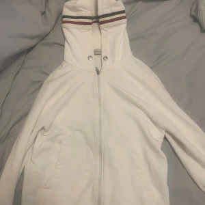Vit hoodie från Moncler med huva - Vit hoodie från Moncler med hel dragkedja, huva och två fickor framtill. Huva med snygga ränder i rött och marinblått. Moncler-logga på ärmen. Tillverkad i mjuk bomull, perfekt för chill dagar. Klassisk och stilren design som funkar året runt.