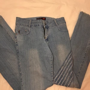 Ljusblå jeans med broderade detaljer - Säljer ett par ljusblå jeans från Teli med snygga broderade ränder på benen och bakfickorna. Klassisk femficksmodell med dragkedja och knapp framtill. Jeansen är bootcut. Viktig att notera att jag köpt dessa begagnade och har slitningar på hällarna och en lös knapp vig gylfen, men inget som syns eller störs. De är ganska långa i benen. Skulle passa någon som är 170+ 