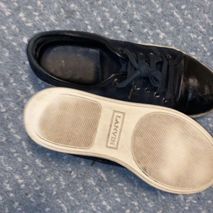 Svarta sneakers från Lanvin i mocka - Snygga svarta sneakers från Lanvin med vit platt sula och rund tå. Ovandelen är i mjuk mocka med detaljer i skinn på tån och snörning framtill. Klassisk låg modell med metallöglor på sidan och diskret branding under sulan. Perfekt för dig som gillar stilrena och lyxiga sneakers.