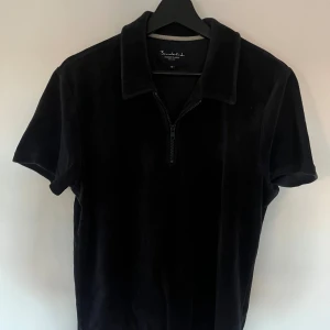 Svart velour t-shirt med krage - Svart t-shirt från Bondelid i mjuk velour med korta ärmar, klassisk krage och half zip-dragkedja framtill. Perfekt för dig som gillar en chill och stilren look med lite retrovibe.