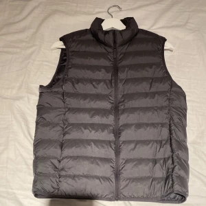 Grå dunväst från Uniqlo XS - Snygg grå dunväst från Uniqlo Ultra Light Down i storlek XS. Västen har en hög krage, horisontella sömmar och dragkedja framtill. Perfekt för lager-på-lager och enkel att bära tack vare sin lätta design.