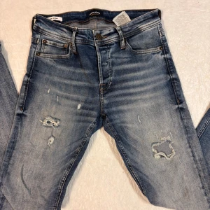 Blå slitna jeans från Jack & Jones - Snygga blå jeans från Jack & Jones med slitningar och hål på framsidan för en cool, avslappnad look. Klassisk femficksmodell med bruna sömmar och läderpatch bak i midjan. Jeansen har slim fit och är tillverkade i mjukt denimtyg.