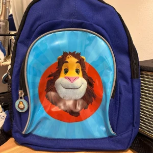 Blå ryggsäck med lejonmotiv - Blå ryggsäck med ett stort, färgglatt lejonansikte på framsidan mot en turkos bakgrund och orange cirkel. Väskan har justerbara svarta axelremmar, dragkedja och ett matchande lejonmotiv på dragkedjans hänge. Perfekt för att sticka ut i skolan eller på utflykten.