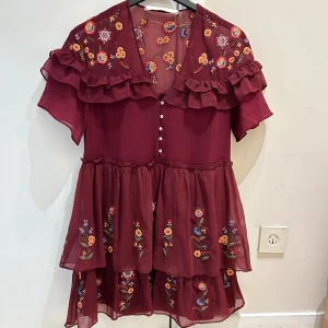  Ny supersöt  Vinröd blommig kortklänning från Zara - Supersöt vinröd kortklänning från Zara med broderade blommor i flera färger, volangdetaljer och knappar framtill. Klänningen har korta ärmar och är gjord i ett lätt, transparent polyestertyg med två lager i kjolen för extra flow. Perfekt för dig som gillar romantisk och boho stil.