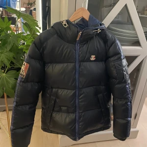 Svart pufferjacka från Dolomite - Snygg svart pufferjacka från Dolomite med broderad röd logga på ryggen och patch på ärmen. Jackan har huva, dragkedja framtill och coola detaljer som snörning vid halsen. Perfekt för kalla dagar och har en sportig vibe.