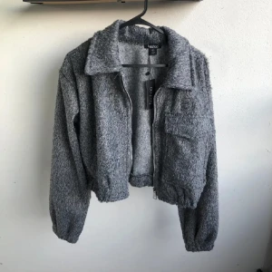 Grå teddyjacka - Mysig grå teddyjacka croppad med fluffig textur, dragkedja framtill och stora ficka på ena sidan med lock. Jackan har en klassisk krage varm o mysig. Den är M men passar S bättre tycker jag