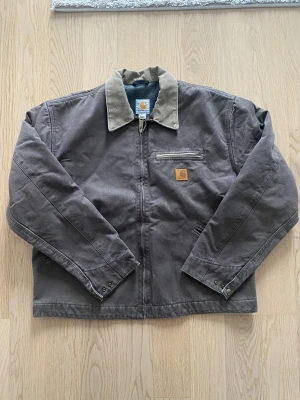 Brun Carhartt workwear jacka M - Klassisk brun workwear jacka från Carhartt i storlek M. Jackan har beige krage, dragkedja framtill, bröstficka med dragkedja och Carhartt-logga. Yttermaterial i kraftig bomull och insida med rutigt foder. Perfekt för dig som gillar streetwear och robust stil.