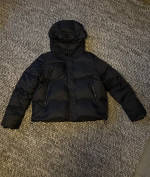 Svart pufferjacka från Snoot - Svart pufferjacka i storlek L, från Snoot Loons med huva och dragkedja framtill. Jackan har två snedställda fickor med dragkedja, justerbar nederkant och en innerficka. Fyllning 90% dun och 10% fjädrar. Dunjacka som är perfekt för kall vinter. Nypris, 4 500kr, men är slutsåld. Helt oanvänd med lapparna kvar. 