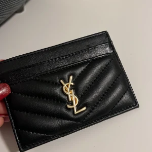 Svart YSL quiltad korthållare i skinn - Supersnygg svart korthållare från YSL i quiltat skinn med guldigt logomärke framtill. Stilren och lyxig design med flera kortfack, perfekt att ha i fickan eller väskan. Klassisk look som passar till allt och känns riktigt exklusiv.