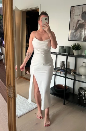 Vit långklänning med slits och smala band - Säljer en elegant vit långklänning med smala axelband som korsas i ryggen. Klänningen har en snygg slits framtill och är gjord i ett glansigt satinliknande material. Perfekt för dig som vill ha en stilren och trendig look.