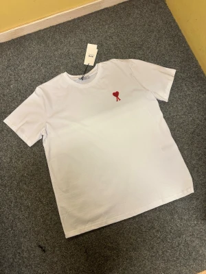 Vit t-shirt från Ami med rött hjärta - Vit t-shirt från Ami i mjuk bomull med klassisk rund hals och korta ärmar. På bröstet finns ett broderat rött hjärta med bokstaven A, vilket ger en clean och stilren vibe. Perfekt för dig som gillar minimalistisk design med en twist.