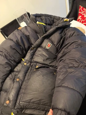 Svart pufferjacka från Fjällräven - Säljer en svart pufferjacka från Fjällräven med gul snörning och tryckknappar i mässing. Jackan har två stora fickor framtill, huva och det klassiska Fjällräven-emblemet på bröstet. Perfekt för kalla dagar och har en riktigt schysst oversized look.har en liten reva vid arm slutet på insidan ingen som syns när man har på den.