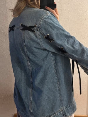 Blå jeansjacka med snörning - Säljer en blå jeansjacka med coola svarta snörningsdetaljer på ryggen och ärmarna. Jackan har klassiska metallknappar och bröstficka med lock. Perfekt för dig som vill ha en unik och trendig denimlook. Den är från gina.