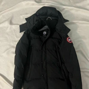 Svart Canada Goose dunjacka med huva - Dragkedjan har lossnat, men man kan fixa det. Storlek S, passar även MSvart dunjacka från Canada Goose med huva och klassisk logopatch på ärmen. Jackan har långa ärmar, stora fickor och knappar framtill. Materialet är slitstark polyester och dunfyllning, perfekt för kalla dagar. Snygg, stilren och riktigt varm.