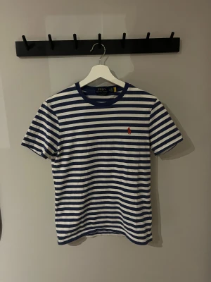 Randig t-shirt från Polo Ralph Lauren - Snygg randig t-shirt från Polo Ralph Lauren i custom slim fit. T-shirten har blå och vita horisontella ränder, På bröstet finns den klassiska röda Polo-loggan. Tillverkad i mjuk bomull för skön känsla.
