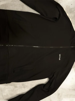 Lyle & Scott svart zip-kofta – herr, storlek XL - Snygg och stilren svart kofta från Lyle & Scott i storlek XL. Modellen har hel dragkedja, diskret broderad logga på bröstet och två sidofickor. Insidan är mjuk och fleecefodrad, vilket gör den både varm och bekväm. Perfekt till vardags, jobb eller lager-på-lager under kallare dagar.  Skick: Bra begagnat skick med normalt litet ludd/fibrer (går lätt att rolla bort). Inga hål eller fläckar. Färg: Svart Detaljer: 	•	Hel dragkedja 	•	Lyle & Scott-logga på bröstet 	•	Mjuk fleeceinsida 	•	Två sidofick