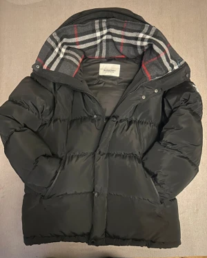 Svart pufferjacka Burberry M - Svart pufferjacka från Burberry i storlek M. Jackan har en klassisk Burberry-rutig huva i grått, vitt och rött, samt dragkedja och knappar framtill. Materialet är polyester och jackan har en loose passform som ger en chill vibe.