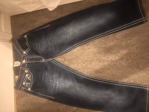 True Religion Billy Super Big T jeans - Säljer ett par mörkblå True Religion Billy Super Big T jeans med kontrasterande vit och orange söm, klassiska fickor med tydlig broderad design och logopatch baktill. Jeansen har normal passform och är tillverkade i robust denim för en cool streetstil.     Jag har tyvärr växt ut de och använt de väldigt väldigt sällan, därför säljer jag de för ett rimligt pris. Det går absolut att diskutera priset vid snabb affär!
