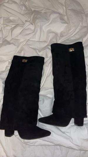 Svarta mockastövlar med klack - Säljer ett par svarta stövlar i mocka med spetsig tå och blockklack. Stövlarna har en snygg guldfärgad detalj upptill och dragkedja på insidan för enkel påtagning. Perfekta för att lyfta din outfit med en stilren och trendig look.