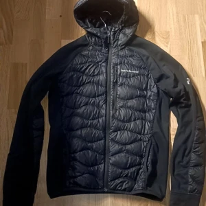 Svart Peak Performance pufferjacka - Svart pufferjacka från Peak Performance med huva och hel dragkedja. Jackan har quiltad framsida och rygg, samt släta ärmar och sidor. Bröstficka med dragkedja och diskret logga på bröstet och ärmen. Perfekt för kyliga dagar. Tagtr mycket väl hand om den i 1 år utan några skado eller defekter den är i bra skick 