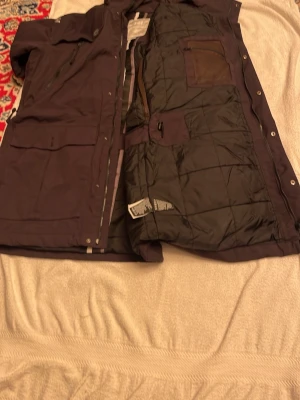 Svart parkajacka Helly Hansen - Svart parkajacka från Helly Hansen med huva, stora fickor framtill och dragkedja samt knappar. Jackan har quiltat foder och flera praktiska innerfickor. Perfekt för kalla dagar och har en clean, funktionell look.