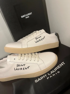 Saint Laurent skor - Snygga vita sneakers från Saint Laurent i skinn med handskriven logga på sidan. Skorna har beige platt sula, snörning och svart detalj på hälen. Perfekta för dig som gillar stilrena och exklusiva sneakers med en twist.