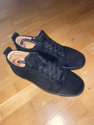 Svarta sneakers från Christian Louboutin - Säljer ett par svarta sneakers från Christian Louboutin i mocka med klassisk röd sula och signaturtext undertill. Skorna har rund tå, snörning och en stilren, minimalistisk design. Perfekta för dig som vill ha något exklusivt och snyggt till din outfit.
