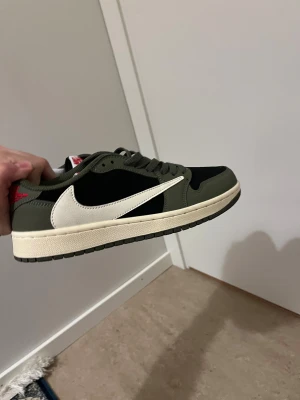 Travis Scott skor  - Nike Air Jordan 1 Low Travis Scott i olivgrönt och svart med vit omvänd swoosh. Röd broderad Air Jordan-logga och Cactus Jack-detaljer på hälen. Ovandel i mocka och skinn, platt sula och snörning. Perfekt för dig som vill sticka ut med en ikonisk sneaker.