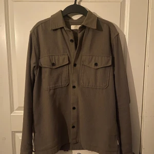 Beige/grön overshirt från Jack & Jones - Stilren beige overshirt från Jack & Jones med klassisk krage, två bröstfickor med lock och svarta knappar framtill. Jackan har lång ärm och är tillverkad i ett mjukt material som passar perfekt för lager-på-lager-stil.