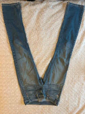 Blå jeans  - Säljer ett par blå jeans från Replay med coola detaljer som förstärkta bakfickor, nitar och justerbara remmar vid midjan. Jeansen har en klassisk femficksdesign och är tillverkade i slitstarkt denim. Perfekt för dig som gillar streetstyle och snygga jeans.