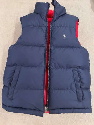 Vändbar dunväst från Polo Ralph Lauren - Snygg vändbar dunväst från Polo Ralph Lauren i barn storlek med marinblå och röd sida. Västen har två fickor på varje sida, dragkedja och ikonisk Polo-logga på bröstet. Fylld med dun och fjädrar för extra värme. Perfekt för att addera färg till din outfit.