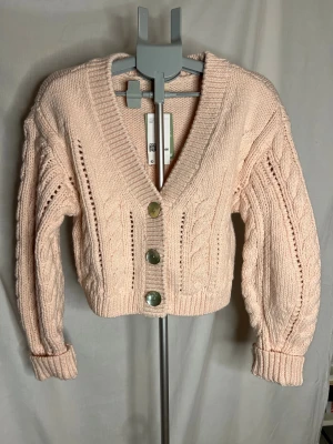 Ljusrosa stickad kofta från H&M - Supermysig ljusrosa stickad kofta från H&M i storlek XS. Koftan har grov kabelstickning, v-ringad hals och tre stora knappar framtill. Modellen är croppad med långa ärmar och breda muddar, perfekt för att ge din outfit en soft vibe.