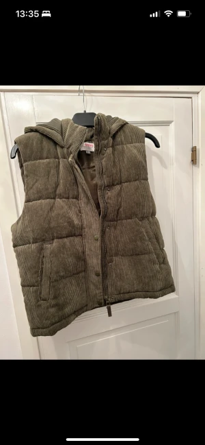 Olivgrön manchester dunväst från CI Sono - Snygg olivgrön dunväst från CI Sono med quiltad design och huva. Västen är tillverkad i mjuk manchester och har praktiska fickor samt både dragkedja och knappar framtill. Perfekt för lager-på-lager och chill stil under höst och vinter.